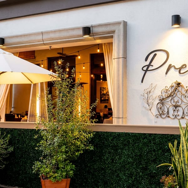 Prego restaurant exterior with open windows, patio umbrella, warm lighting, greenery, and decorative wall signage.
