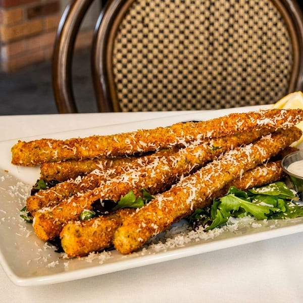 Crispy breaded vegetable sticks topped with grated cheese, served over greens with dipping sauce on a restaurant plate.
