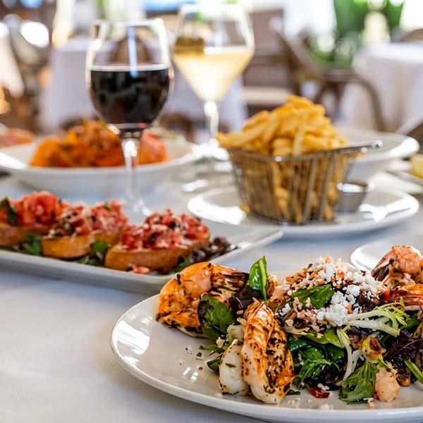 Grilled shrimp with leafy greens and crispy topping sits on a white plate among assorted dishes and wine glasses.