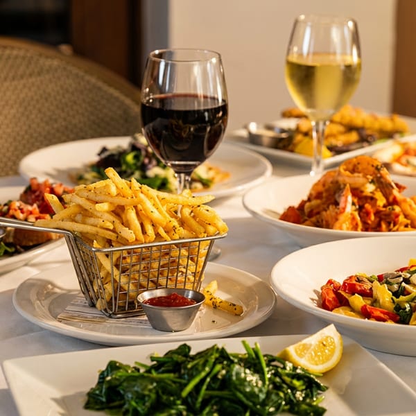 Italian restaurant table featuring pasta dishes, pizza, bruschetta, fries with dipping sauce, spinach, and glasses of red and white wine.