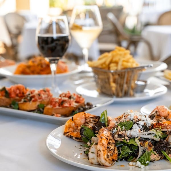 Outdoor restaurant table featuring grilled shrimp salad, fries, assorted dishes, wine, and fresh juice.