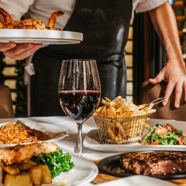 Server presents a plate of shrimp over a restaurant table filled with steak, fries, pasta, salad, wine, and cocktails.
