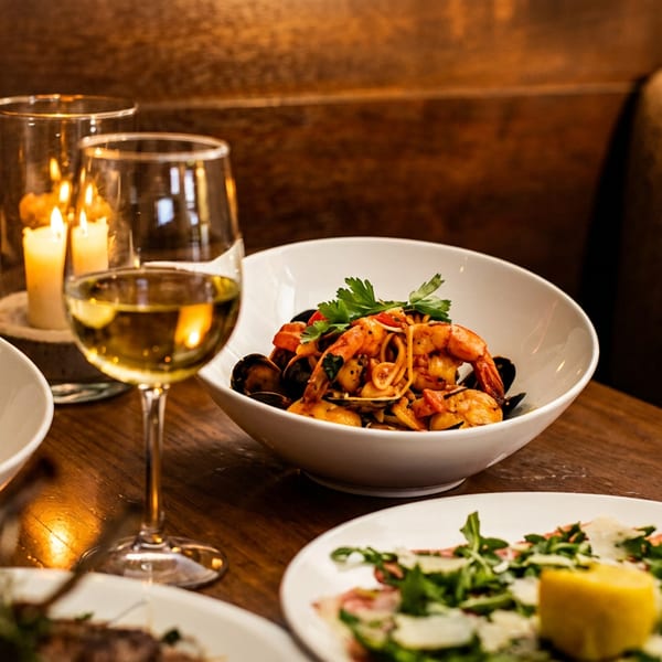 Candlelit restaurant table with seafood pasta, plated dishes, a glass of white wine, and warm upholstered seating.